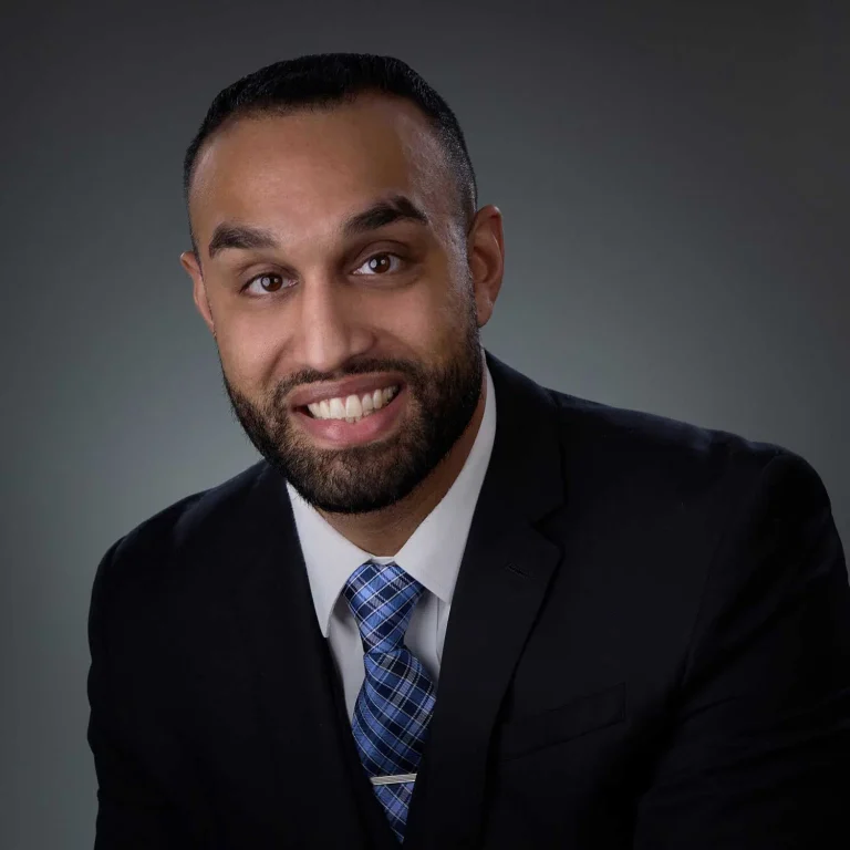 Man with short dark hair and beard, smiling, wearing a black suit jacket, white dress shirt, and blue plaid tie, against a plain gray backdrop.