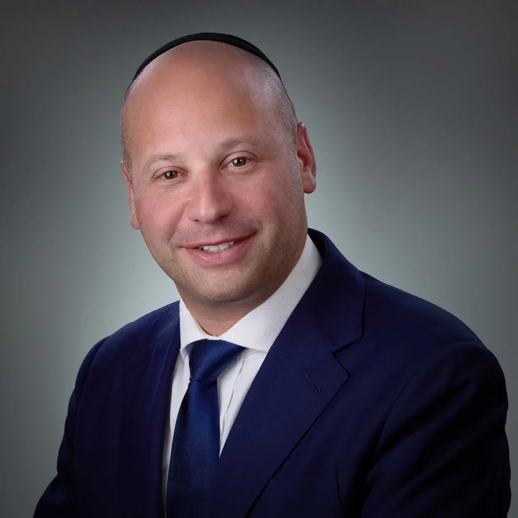 Smiling bald man wearing a kippah, navy blue suit, white shirt, and blue tie against a gray gradient background