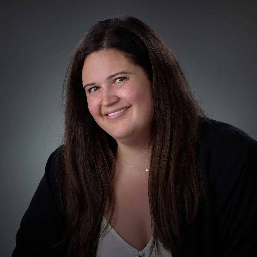 Portrait of a smiling woman with long dark hair, wearing a black jacket over a white top, against a dark gray background.