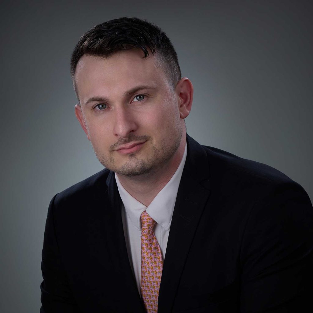 Portrait of a man with short dark hair, wearing a black suit jacket, white shirt, and pink patterned tie, against a plain gray background.