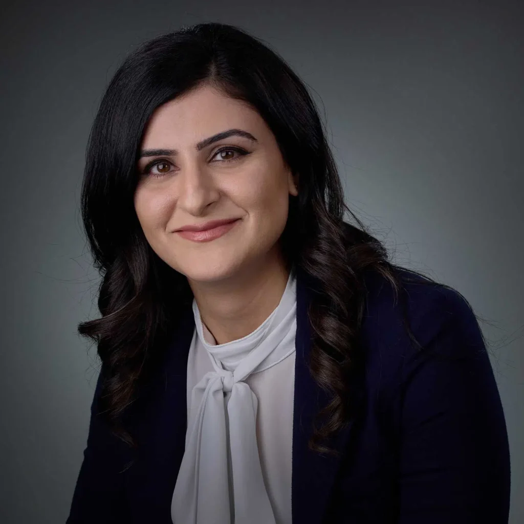 Smiling woman with long black hair, wearing a white blouse with a bow tie and a dark blazer, posing against a gray background.
