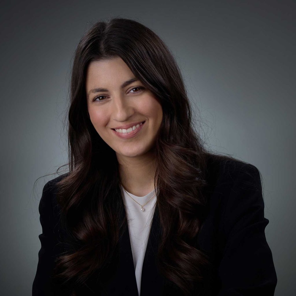Portrait of a smiling woman with long dark wavy hair wearing a black blazer, white top, and a small necklace against a neutral gray background.