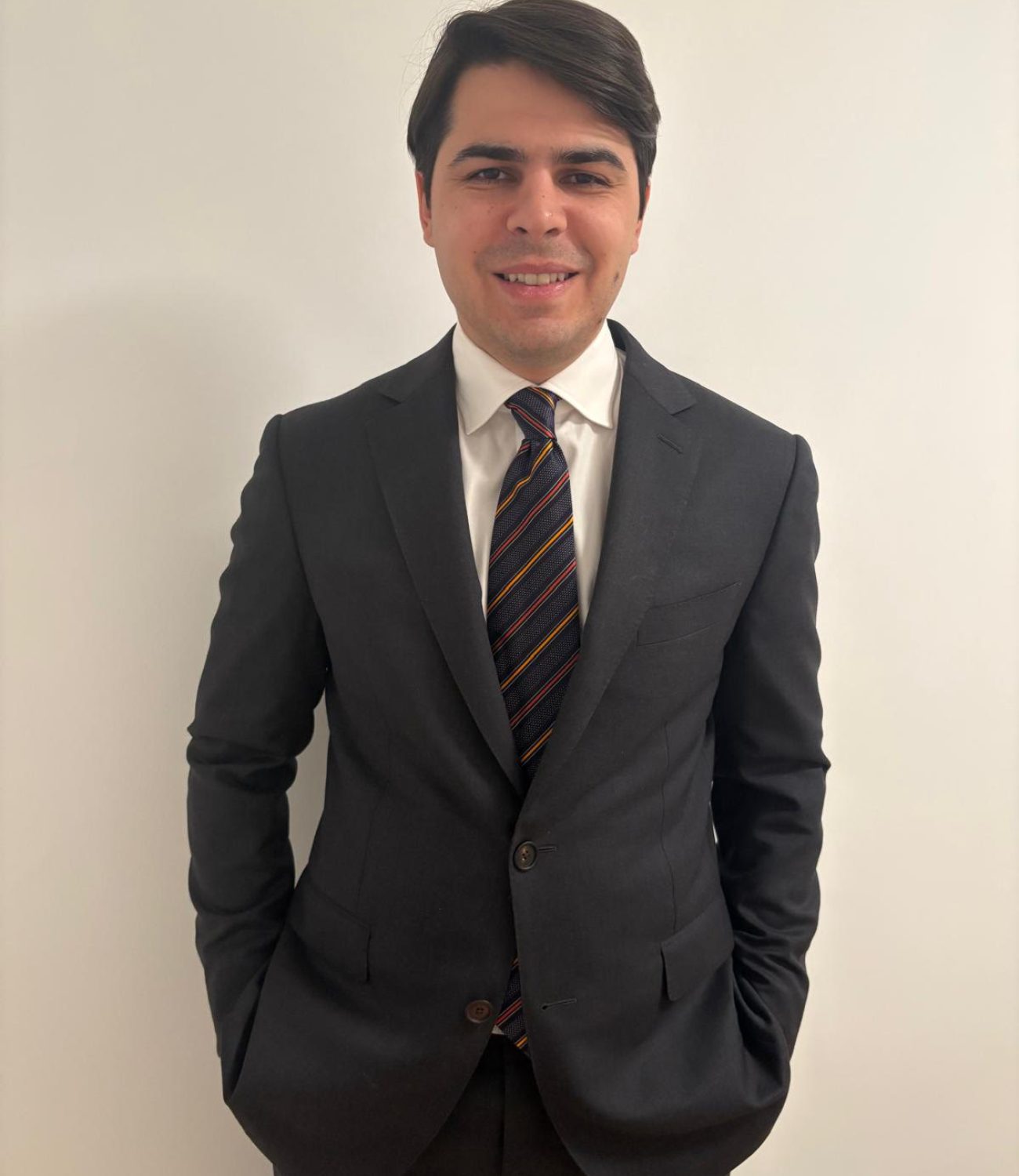 Portrait of a smiling young man with dark hair wearing a black suit, white shirt, and striped tie, standing against a plain white background with his hands in his pockets.