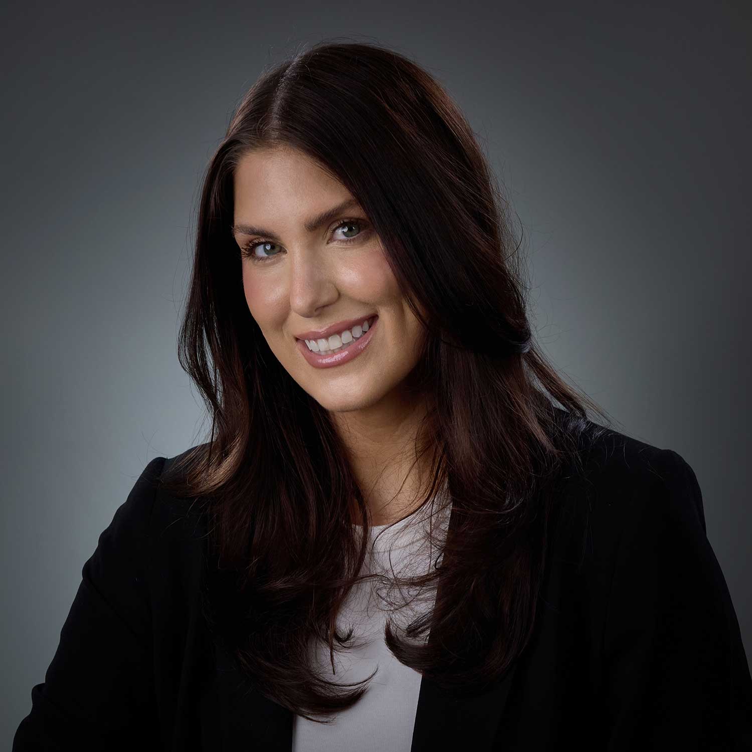 Smiling woman with long dark brown hair wearing a black blazer and white top against a gray background.
