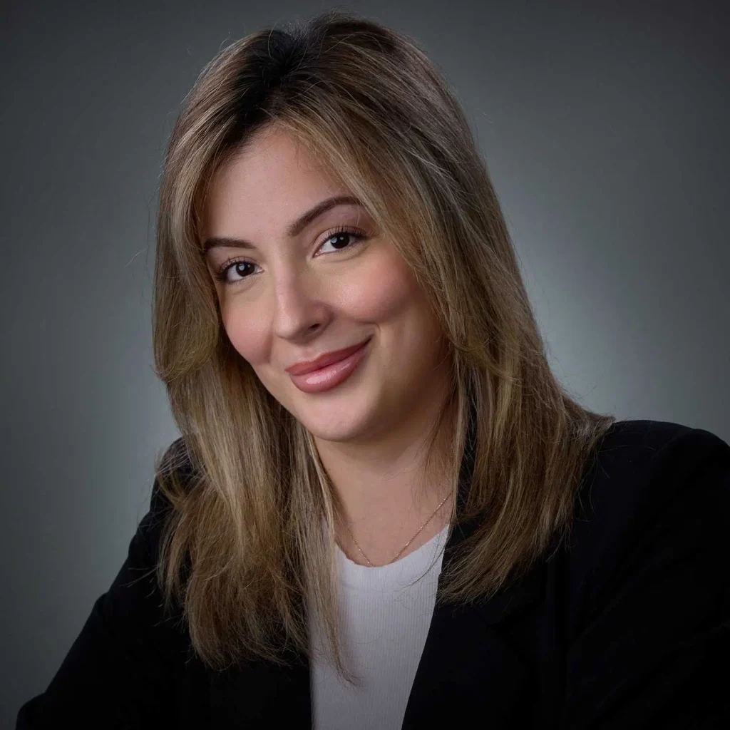 Portrait of a smiling woman with light brown hair, wearing a black blazer and white top, against a plain gray background.