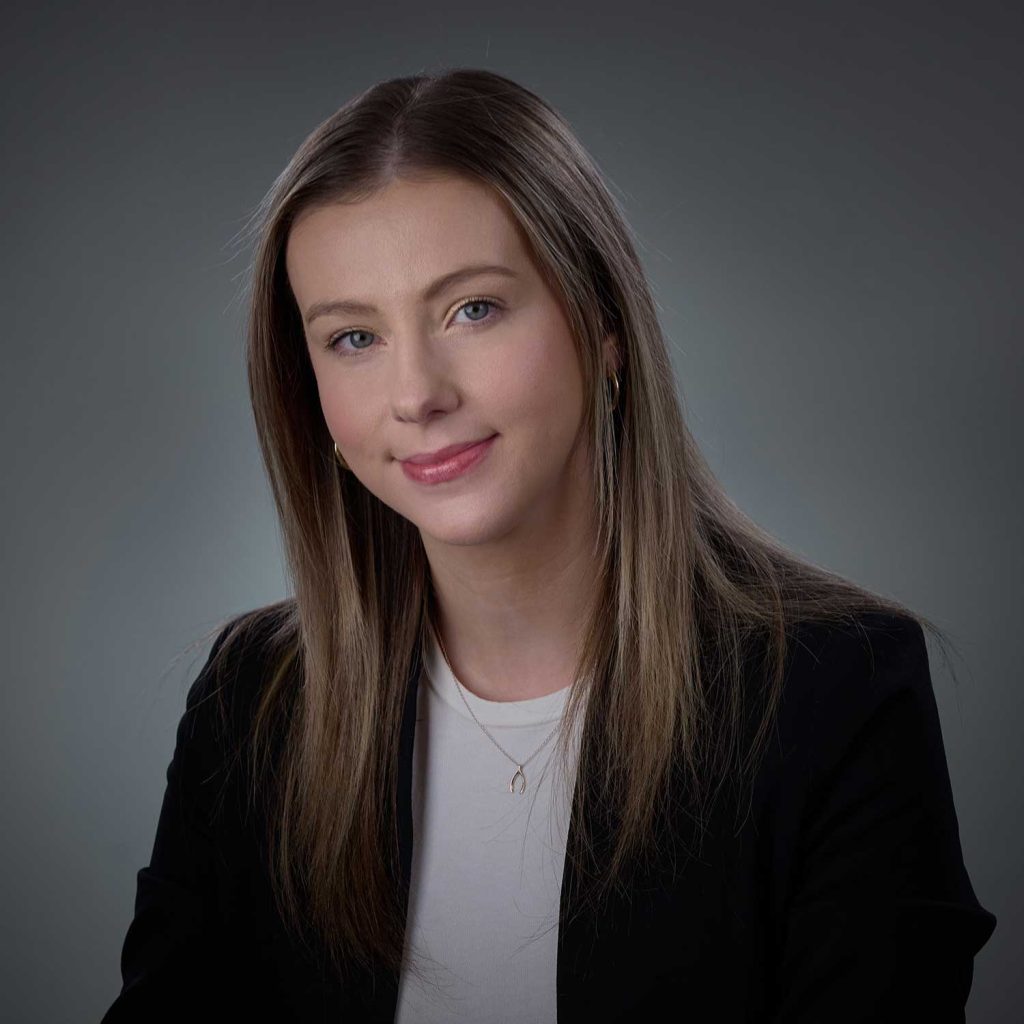 Young woman with straight light brown hair wearing a black blazer, white top, and a necklace with a wishbone pendant, smiling slightly against a plain gray background.