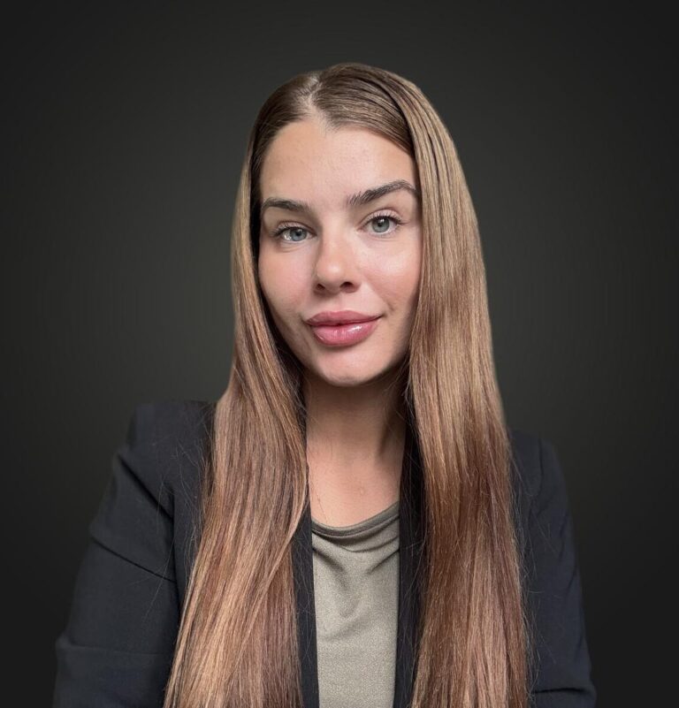 Portrait of a woman with long straight light brown hair, wearing a black blazer and an olive green top, against a dark gray background.