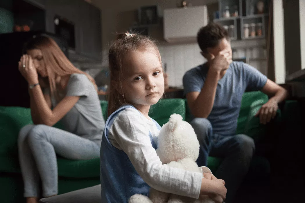 A sad young girl holding a teddy bear sits in the foreground while her distressed parents sit apart on a couch, symbolizing family conflict and the need for family legal support in Toronto.