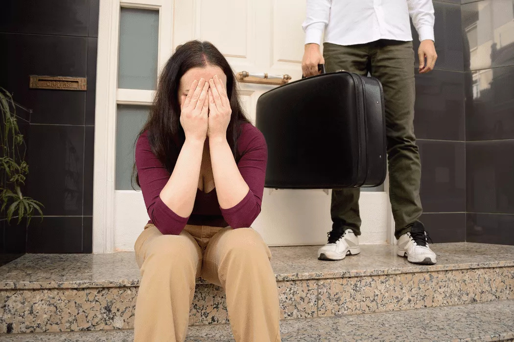 A distressed woman sitting on the steps outside a building covering her face with her hands while a man stands behind her holding a suitcase, symbolizing family separation and legal struggles addressed by Nussbaum family lawyers in Toronto.