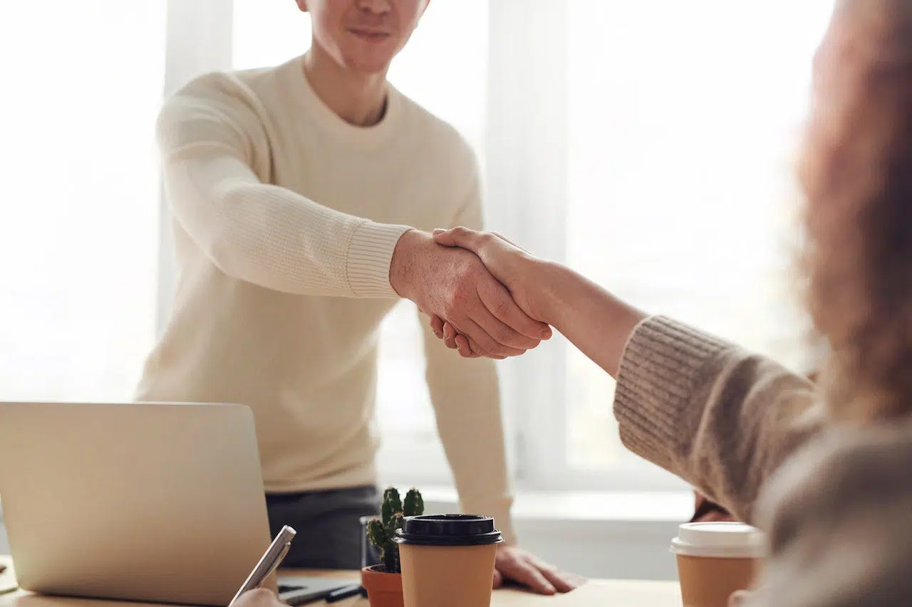 Two people shaking hands in a bright office setting with a laptop, coffee cups, and a small plant on the table, symbolizing a professional agreement or consultation, representing Nussbaum family lawyers in Toronto.