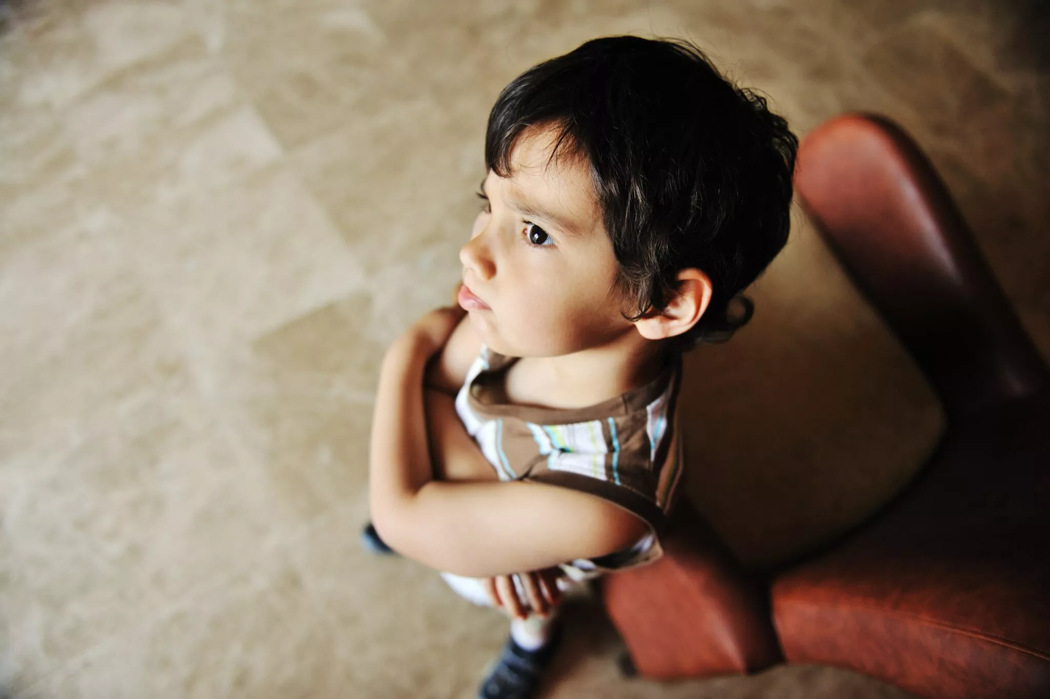 A young child standing with arms crossed, looking up pensively beside a brown chair, representing the emotional impact often seen in family law cases handled by Nussbaum Family Lawyers in Toronto.